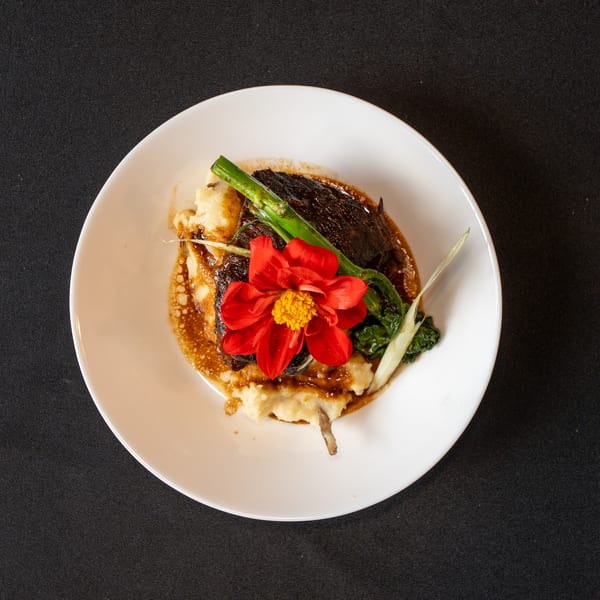 Braised short rib with mashed potatoes, green vegetables, and a red flower garnish on a white plate.