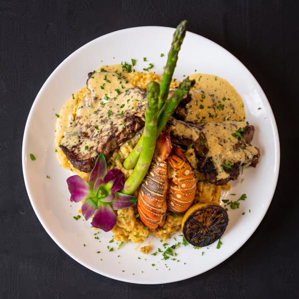 Steak and lobster tails with asparagus, grilled lemon, and a purple orchid on a white plate.