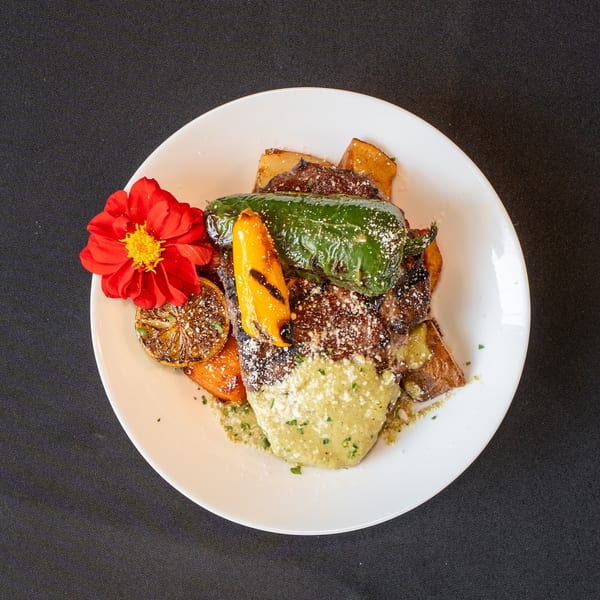 Grilled steak with green sauce, roasted peppers, potatoes, and a red flower garnish on a white plate.