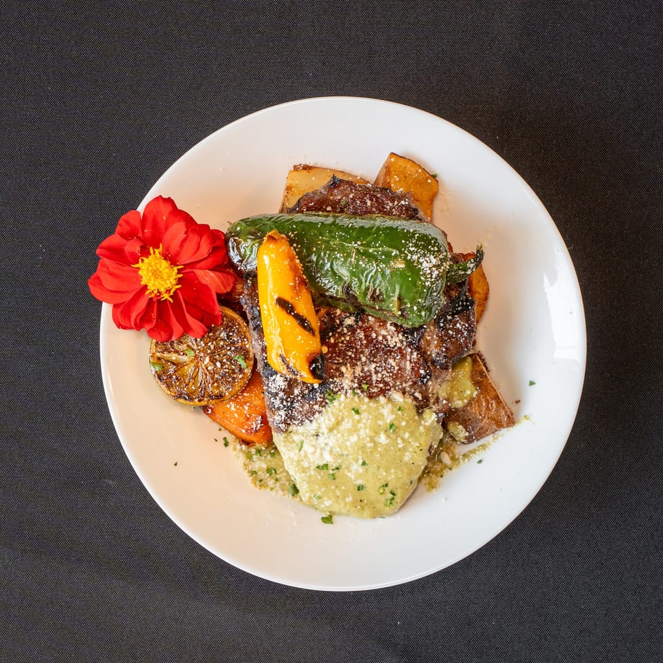 Grilled steak with green sauce, roasted peppers, potatoes, and a red flower garnish on a white plate.