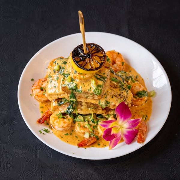 Seafood dish with shrimp, fish, creamy sauce, charred lemon, and an orchid on a white plate.