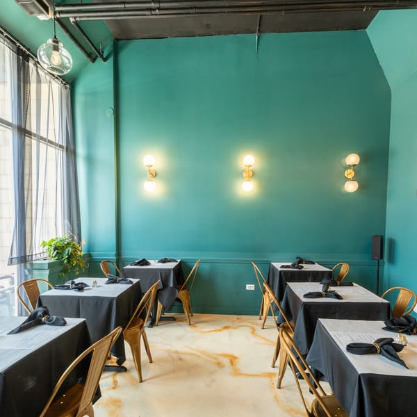 Restaurant dining room with teal walls, black-draped tables, gold chairs, and large windows.