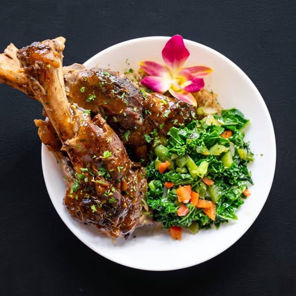 Braised lamb shanks with gravy, green vegetables, and a pink orchid in a white bowl.