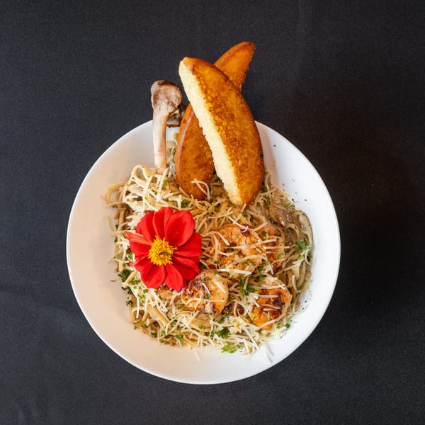 Shrimp pasta with grated cheese, parsley, garlic bread, and a red flower garnish in a white bowl.