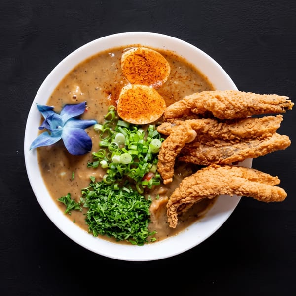 A bowl of gumbo with fried fish, hard-boiled eggs, green onions, and parsley, garnished with a blue orchid.