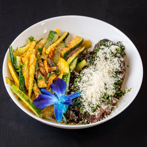 A white bowl filled with steak, zucchini, squash, and a blue orchid garnish on a dark surface.