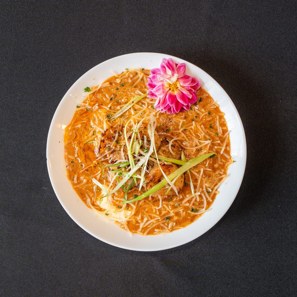 Overhead view of a white bowl filled with noodles, shrimp, and a pink flower garnish.