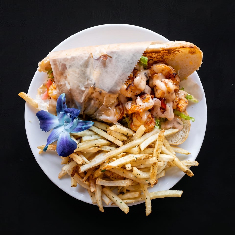 Shrimp po'boy sandwich with fries and a blue orchid on a white plate.