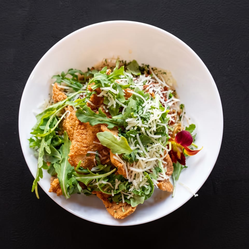 Crispy fried chicken, arugula, and shredded cheese in a white bowl on a dark surface.