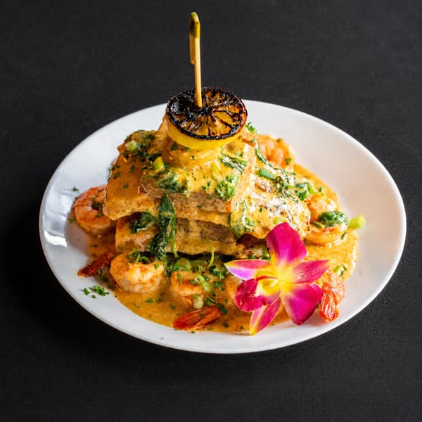 Stacked fish fillets with shrimp, creamy sauce, grilled lemon, and an orchid on a white plate.