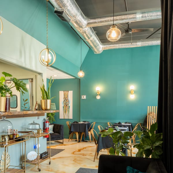 A modern restaurant interior with teal walls, gold accents, black tables, and lush green plants.