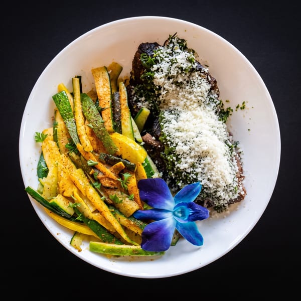 Grilled fish with parmesan, herbs, and roasted zucchini and squash, garnished with a blue orchid.