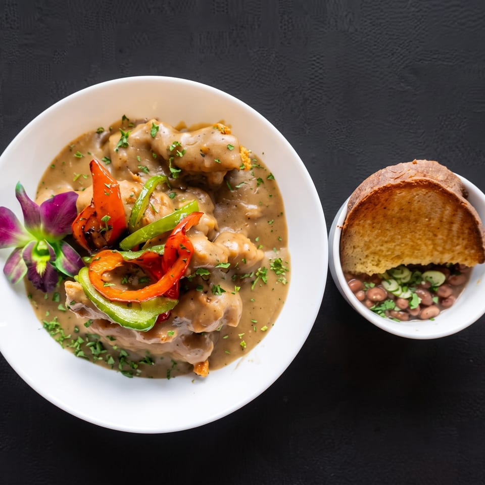 Chicken and gravy with bell peppers and orchids, alongside a bowl of beans and toast.