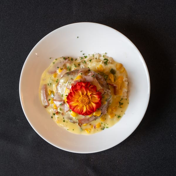 Savory dish with a meat patty, creamy sauce, corn, rice, and a red-orange flower garnish.