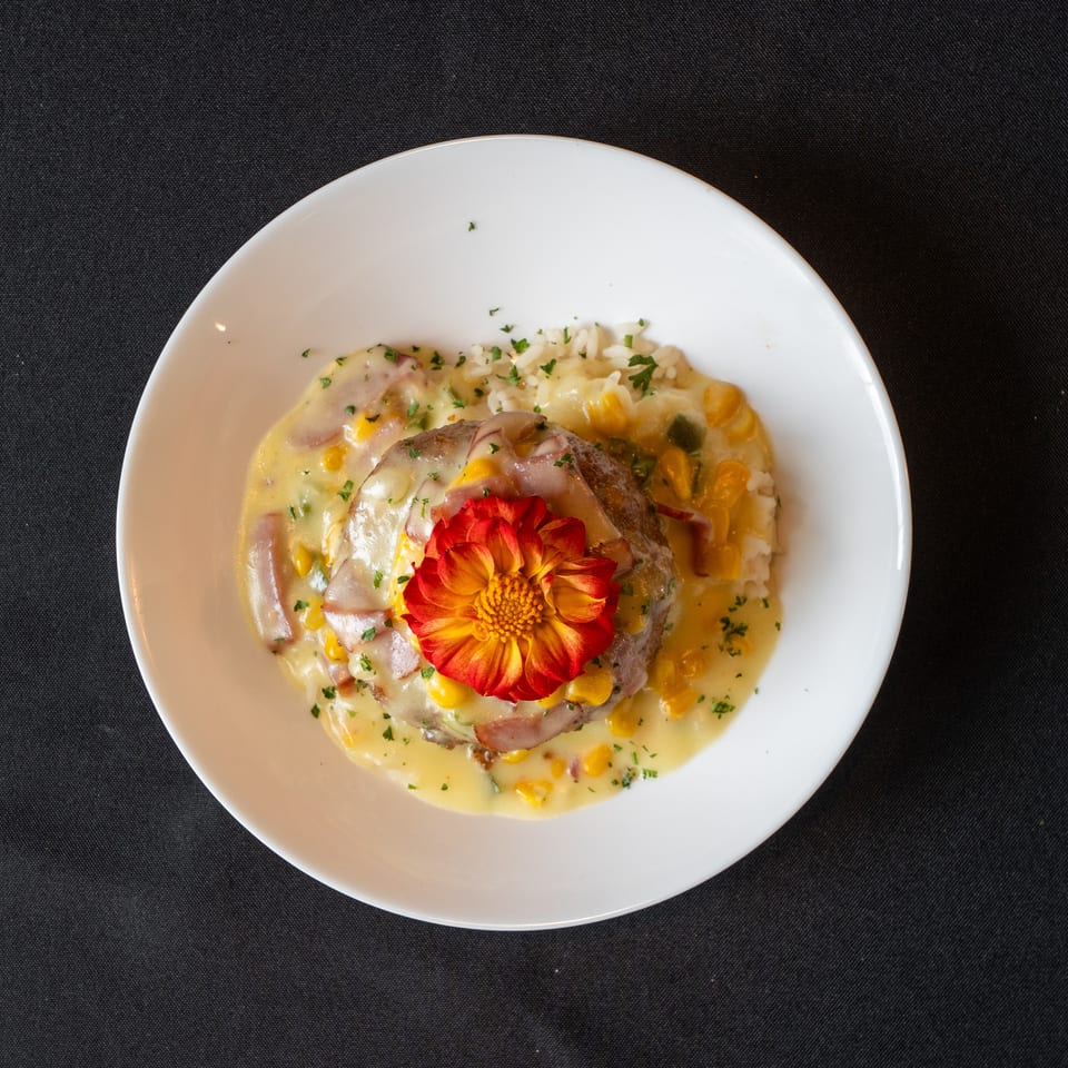 Savory dish with a meat patty, creamy sauce, corn, rice, and a red-orange flower garnish.